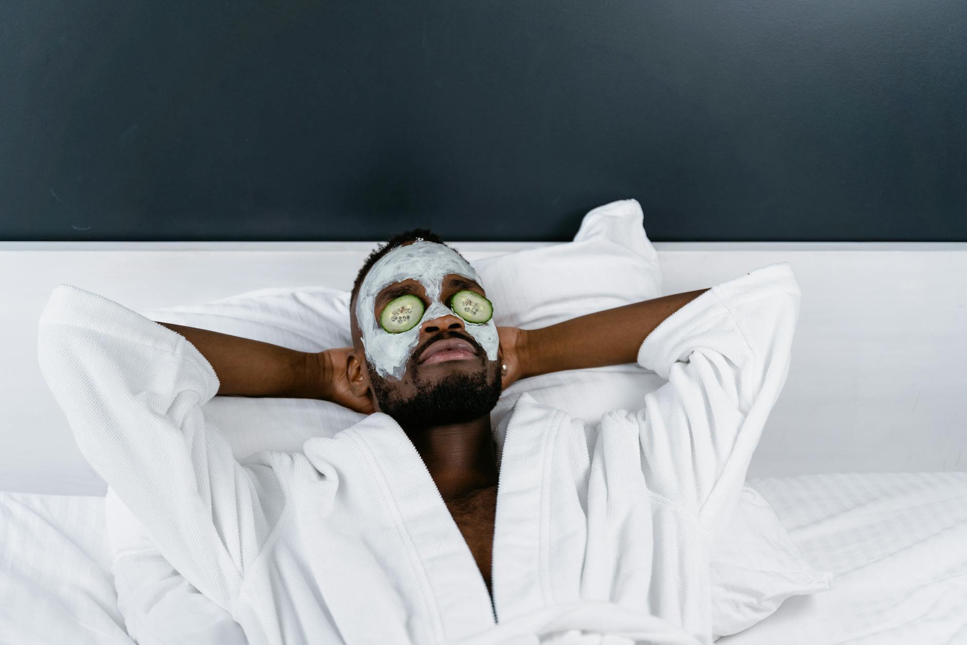 A man relaxing on a bed wearing a white bathrobe, with a green facial mask and cucumber slices over his eyes.
